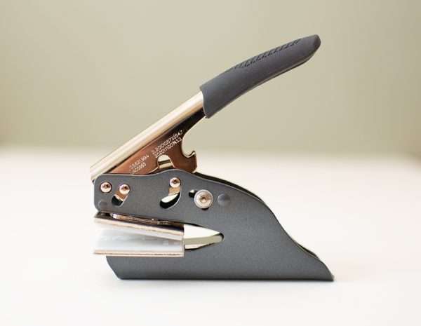 UK handheld embosser with rectangular die plate on beige background.